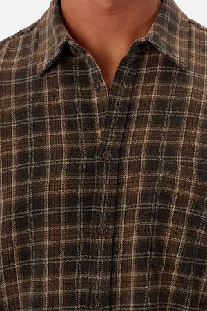 A close-up of a person wearing the Industrie The Maple Long Sleeve Shirt, featuring a brown and black cotton checked pattern, collar, visible buttons, and long sleeves. Only the lower face, neck, and upper torso are shown.