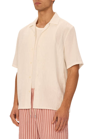 A man models the Industrie The Marbella Short Sleeve Shirt in cream over a white tee, paired with pink and white striped shorts for a laid-back everyday look against a plain backdrop.
