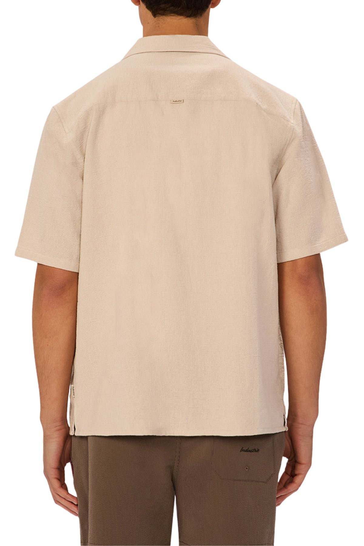 A person is seen from behind wearing the Industrie The Marbella Short Sleeve Shirt in light beige, made from lightweight textured fabric with a spread collar and short sleeves, paired with brown pants against a plain white background.