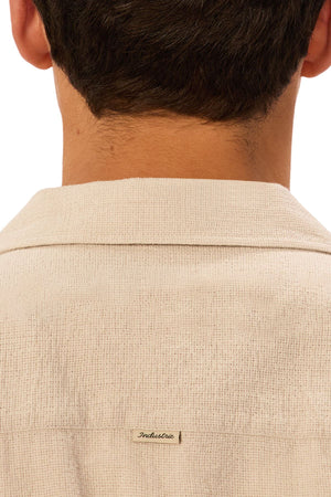 Close-up of the back of a person's neck and shoulders wearing the Industrie The Marbella Short Sleeve Shirt in cream textured fabric. A small rectangular "Industrie" tag is sewn below the collar of this relaxed fit shirt.