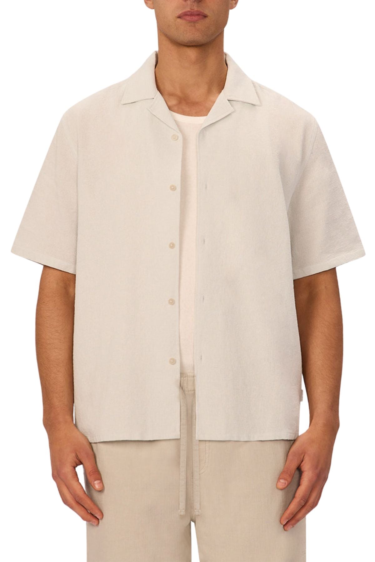 A man wears the Industrie The Marbella Short Sleeve Shirt—a relaxed fit, lightweight textured shirt in light beige—over a white tee with matching beige drawstring pants, standing against a white background.