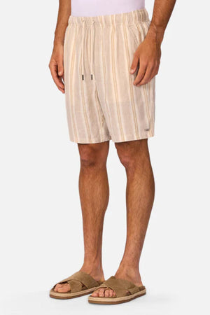 A man wears Industrie The Matias Short—beige and white striped linen shorts—in breathable fabric with tan slide sandals, standing against a light gray background. Only his body from mid-torso to feet is shown, highlighting casual style.