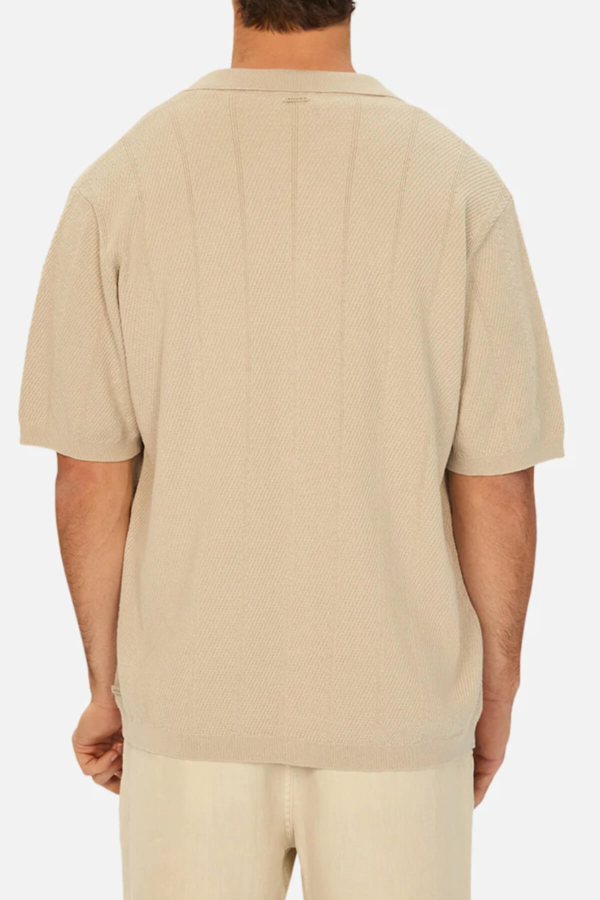 From the back, someone wears a loose-fitting Industrie The Modena Short Sleeve Shirt. This beige polo features a camp collar and knitted texture, presenting a subtle vertical pattern with short sleeves against a plain white background, highlighting only the upper body.