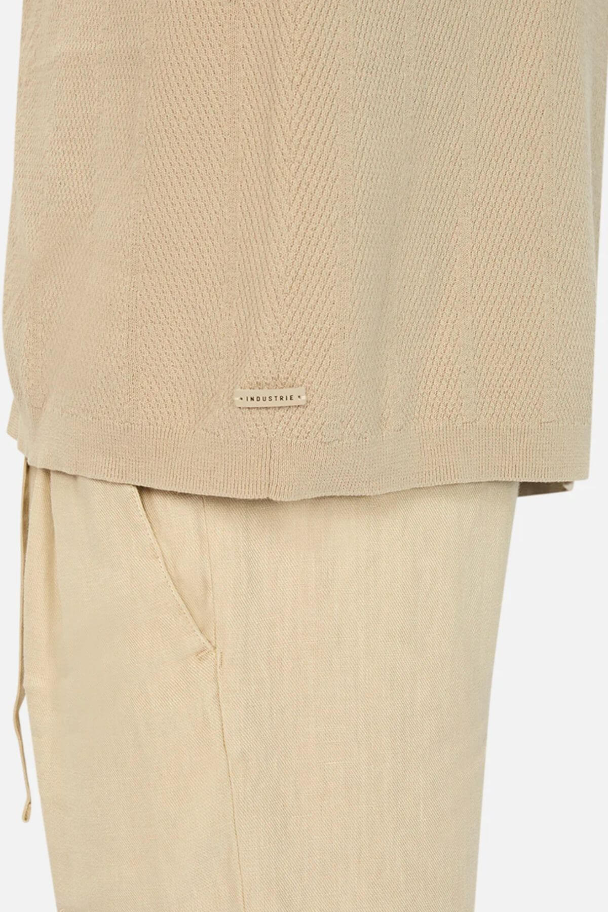 Close-up of a person in the Industrie The Modena Short Sleeve Shirt, a beige textured design with a camp collar, paired with matching pants. The shirt displays an "INDUSTRIE" label. Pants feature visible side pocket and drawstring, set against a plain white background.