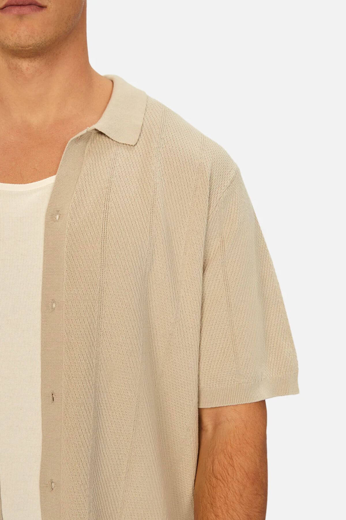 A person is wearing the Industrie The Modena Short Sleeve Shirt, which features a knitted texture and camp collar in beige, layered over a white tank top. Only the upper torso and part of the face are visible against a plain background.
