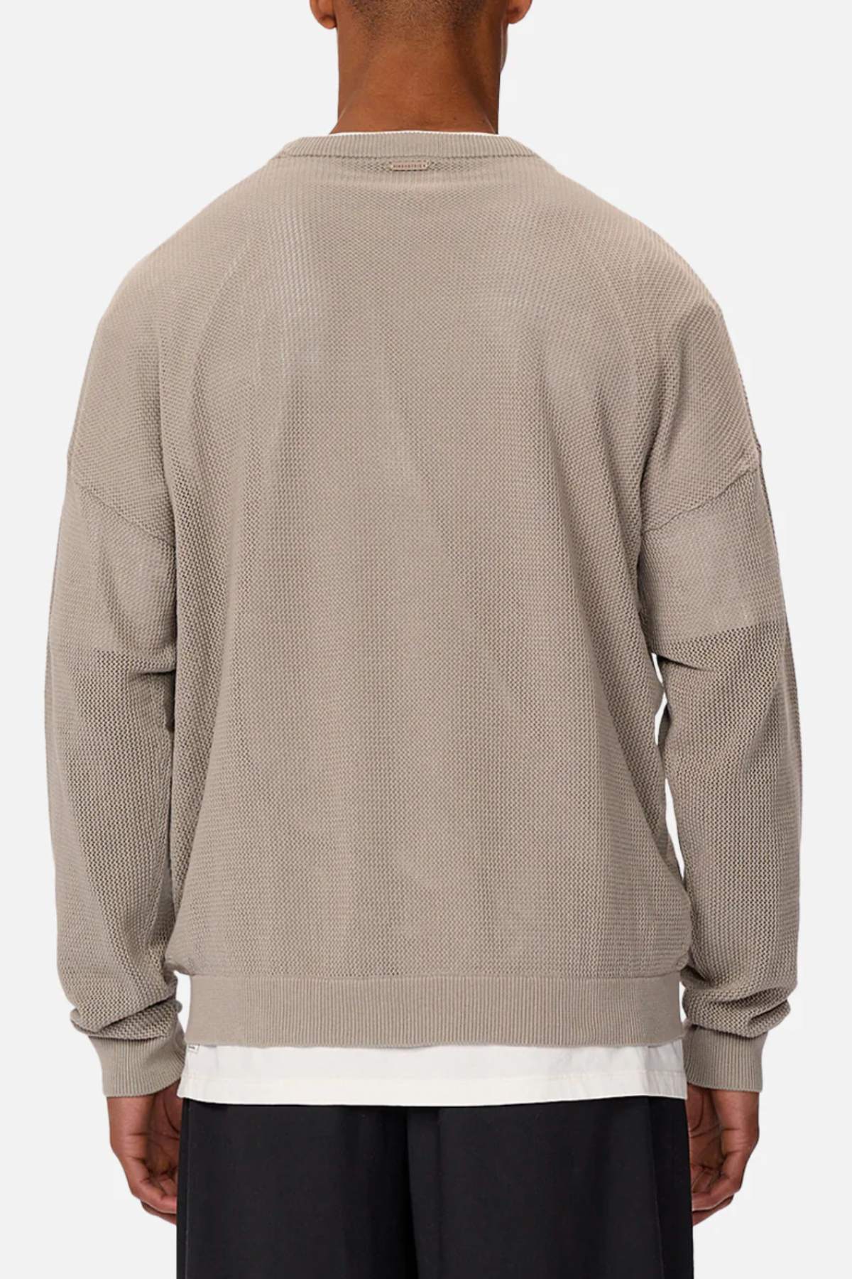 Someone is wearing Industrie The Nanterre Crew Neck Knit, showcasing an oversized fit and textured beige fabric with dropped shoulders. The long sleeves reveal a white shirt underneath, paired with dark pants against a plain backdrop for a stylish look.