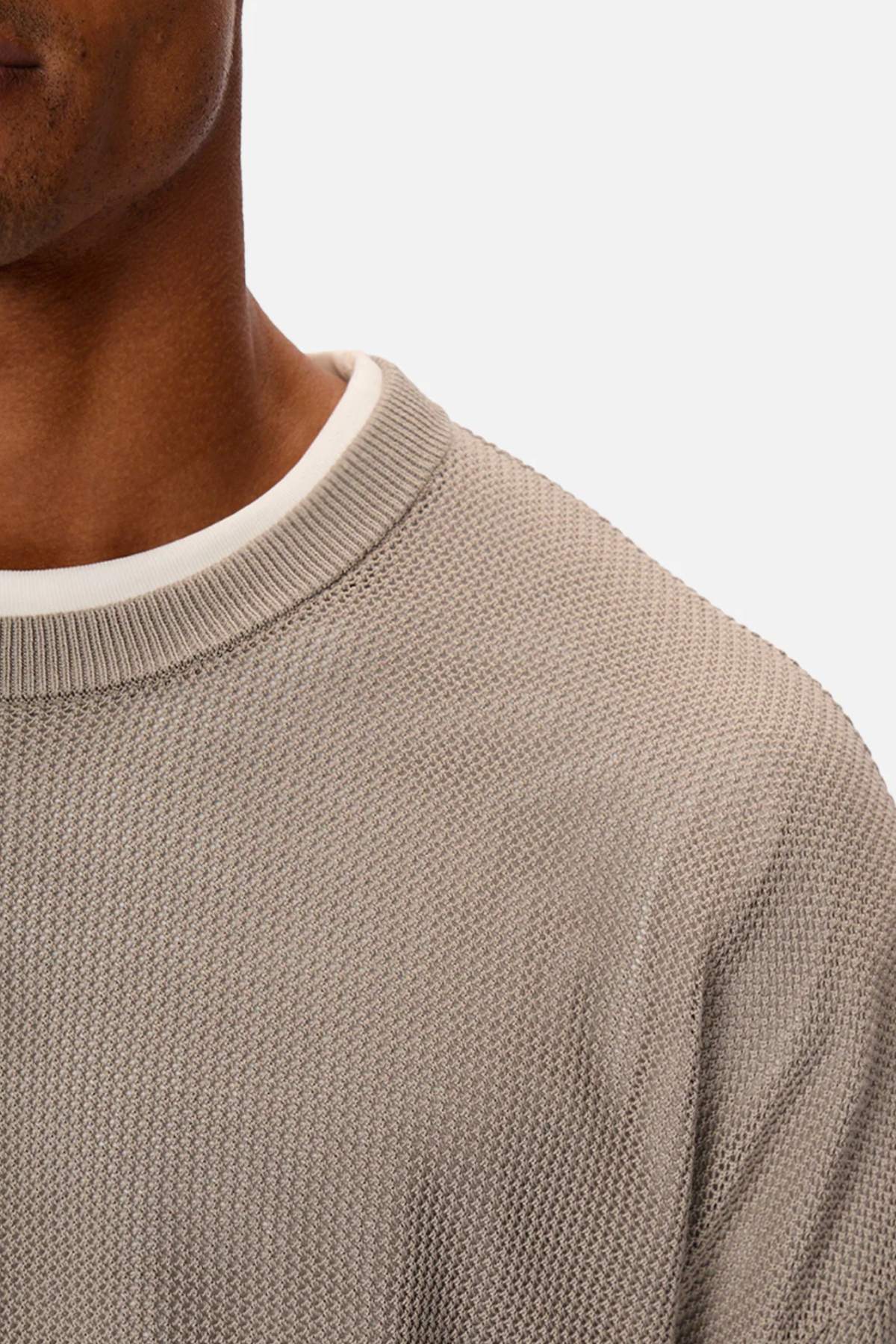 A person partially visible wears Industrie’s The Nanterre Crew Neck Knit, a textured grey oversized sweater made of 100% cotton, over a white round-neck shirt. The focus is on the shoulder and upper chest against a plain light background.