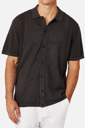 A man wears the Industrie The Nanterre Short Sleeve Shirt, a black button-up with a camp collar and textured pattern, paired with white pants. His left hand in his pocket highlights an effortlessly casual look.
