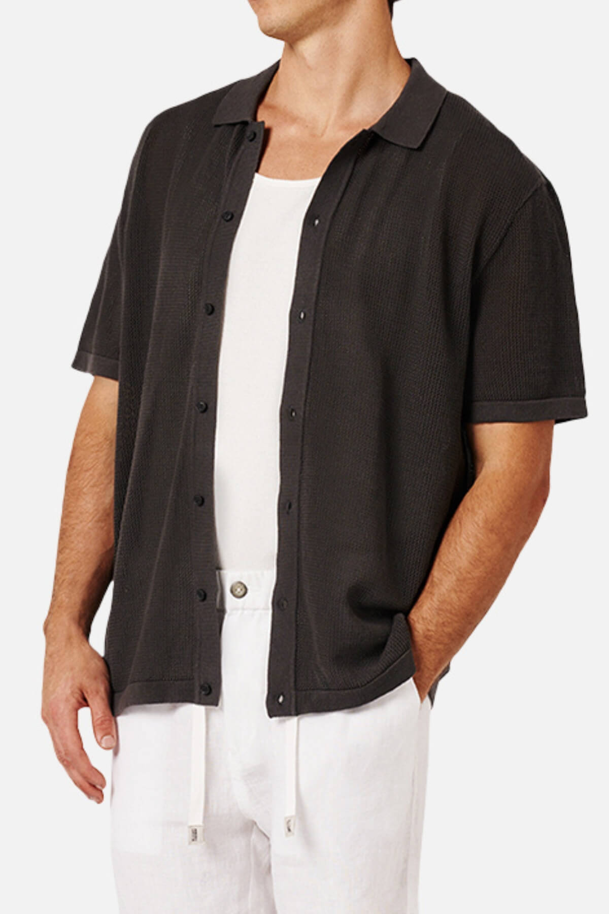 A man wears the Industrie The Nanterre Short Sleeve Shirt in black over a white tank, paired with white drawstring pants. His left hand is in his pocket and his face isn't visible, showcasing a relaxed, casual look.