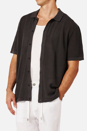 A man wears the Industrie The Nanterre Short Sleeve Shirt in black over a white tank, paired with white drawstring pants. His left hand is in his pocket and his face isn't visible, showcasing a relaxed, casual look.