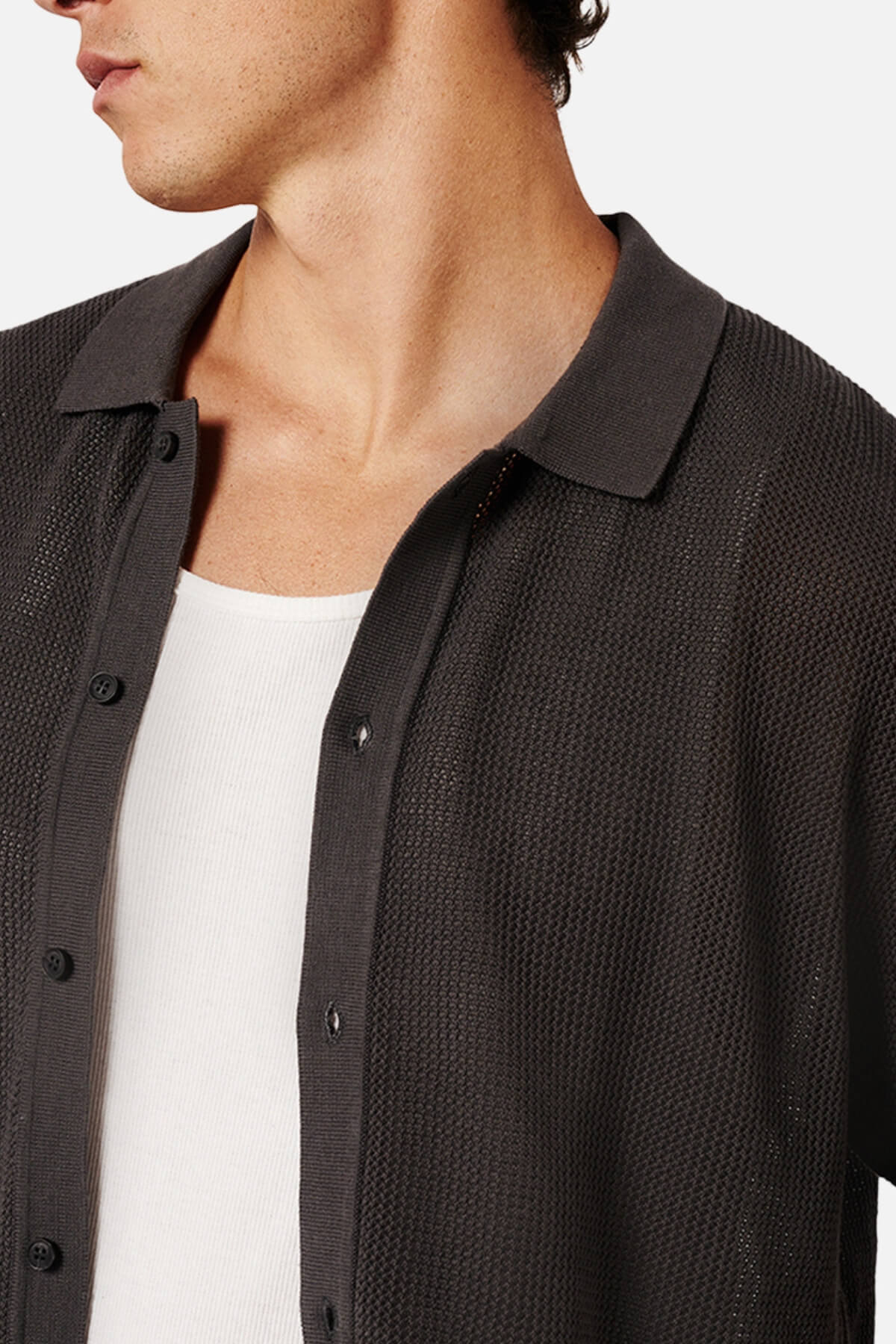 A man wears the Industrie The Nanterre Short Sleeve Shirt, a textured dark button-up with a camp collar, over a white ribbed tank top. Only his chin to mid-torso is visible against a plain background.