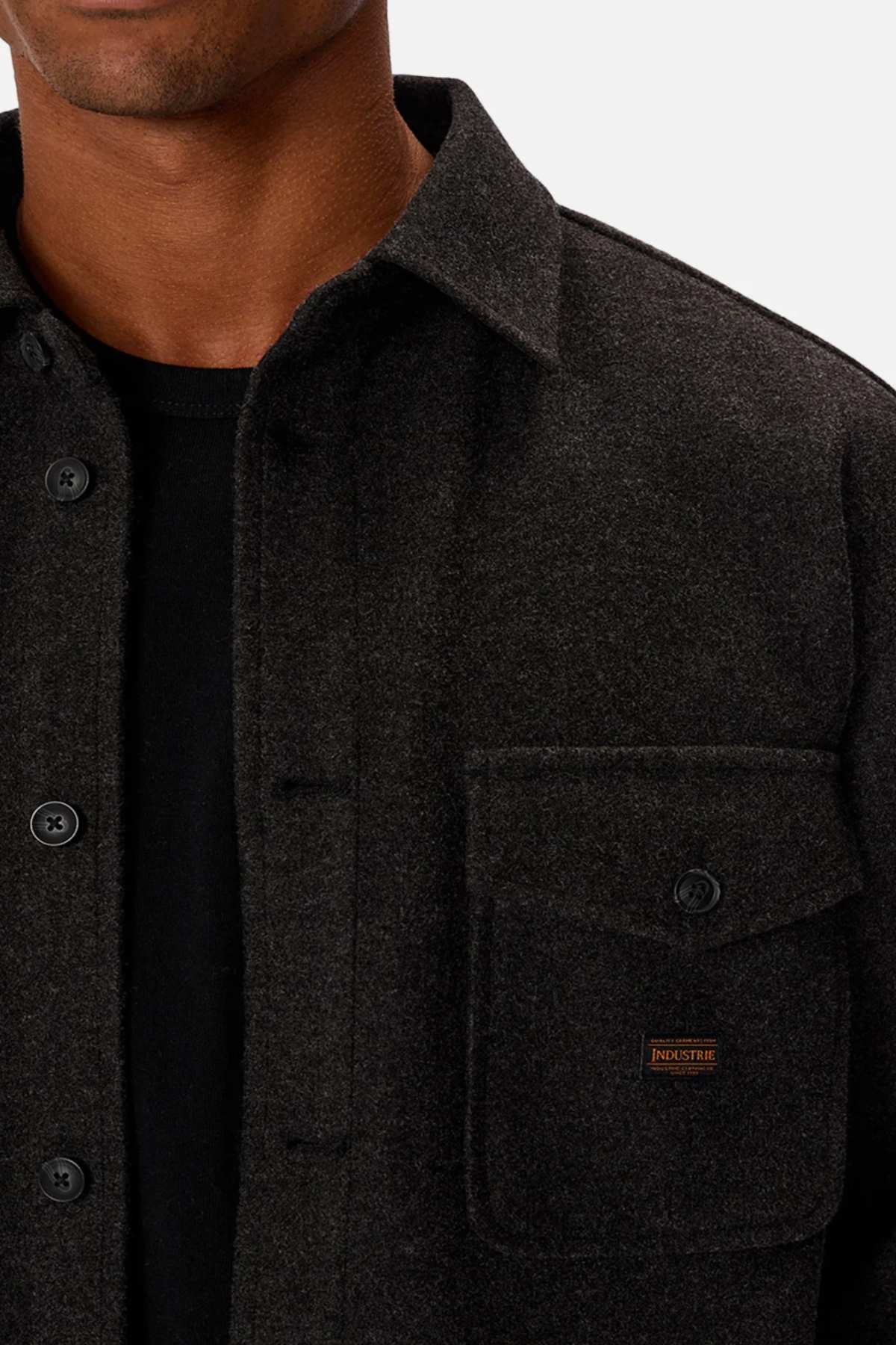 Someone is wearing Industrie The New Coleman Jacket, which has a relaxed fit and classic collar, over a black shirt. The dark gray jacket features a visible chest pocket with a small stitched label.
