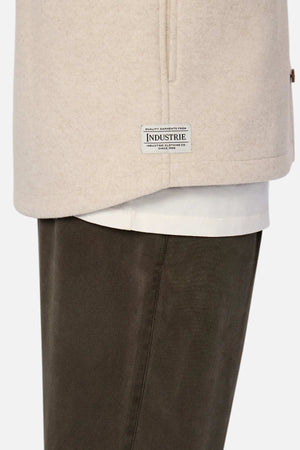 A close-up side view shows a person in the Industrie The New Coleman Jacket—a relaxed-fit beige coat worn over a white shirt with dark brown pants. A rectangular "INDUSTRIE" label near the hem adds a classic touch.