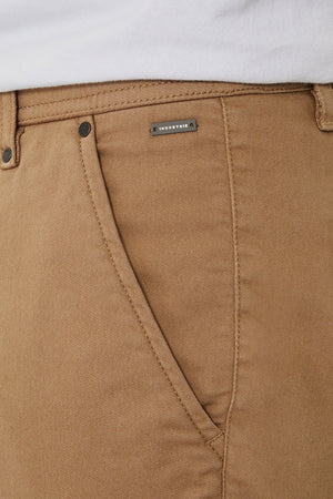 Close-up of Industrie The New Drifter Cuba Short in tan, featuring a side pocket, metal rivets, and a rectangular "INDUSTRIE" label below the waistband. The edge of a white shirt at the top highlights these casual, stylish shorts.