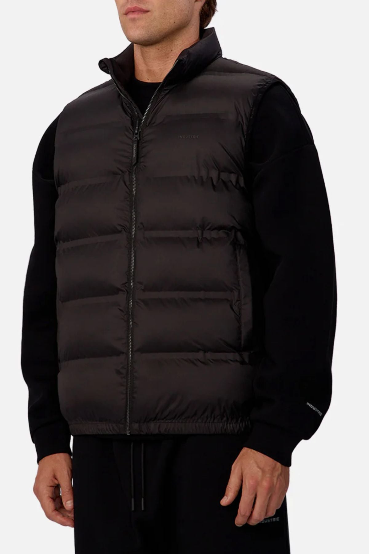 Someone stands against a plain light background wearing the Industrie The New Kyoto Puffer Vest—zipped up over a black long-sleeve shirt and black pants. The vest features heat-sealed seams and a high collar.