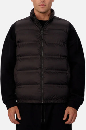 Someone stands facing forward against a plain light background, wearing the Industrie The New Kyoto Puffer Vest over a black long-sleeve shirt. The vest features horizontal quilting, heat-sealed seams, and a sleek front zipper.