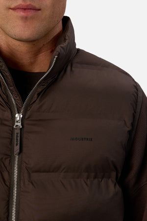 Close-up of a person wearing the Industrie The New Kyoto Puffer Vest, featuring a featherlight design, "INDUSTRIE" chest print, visible zipper, and cozy build—ideal for cooler weather against a plain, light background.