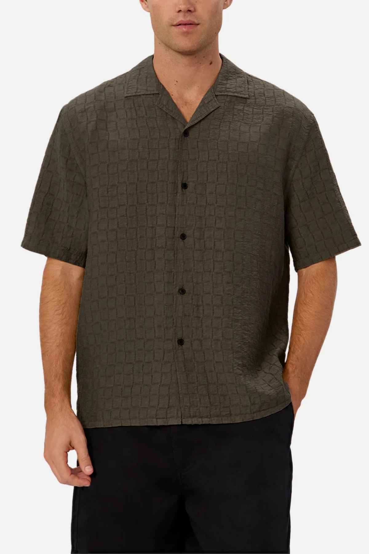 A man is sporting the Industrie The New Ortigia Short Sleeve Shirt, which showcases a dark green textured design with a button-up front and a subtle jacquard pattern. This stylish shirt features a camp collar. He pairs it with black pants, casually placing one hand in his pocket against a plain white background.