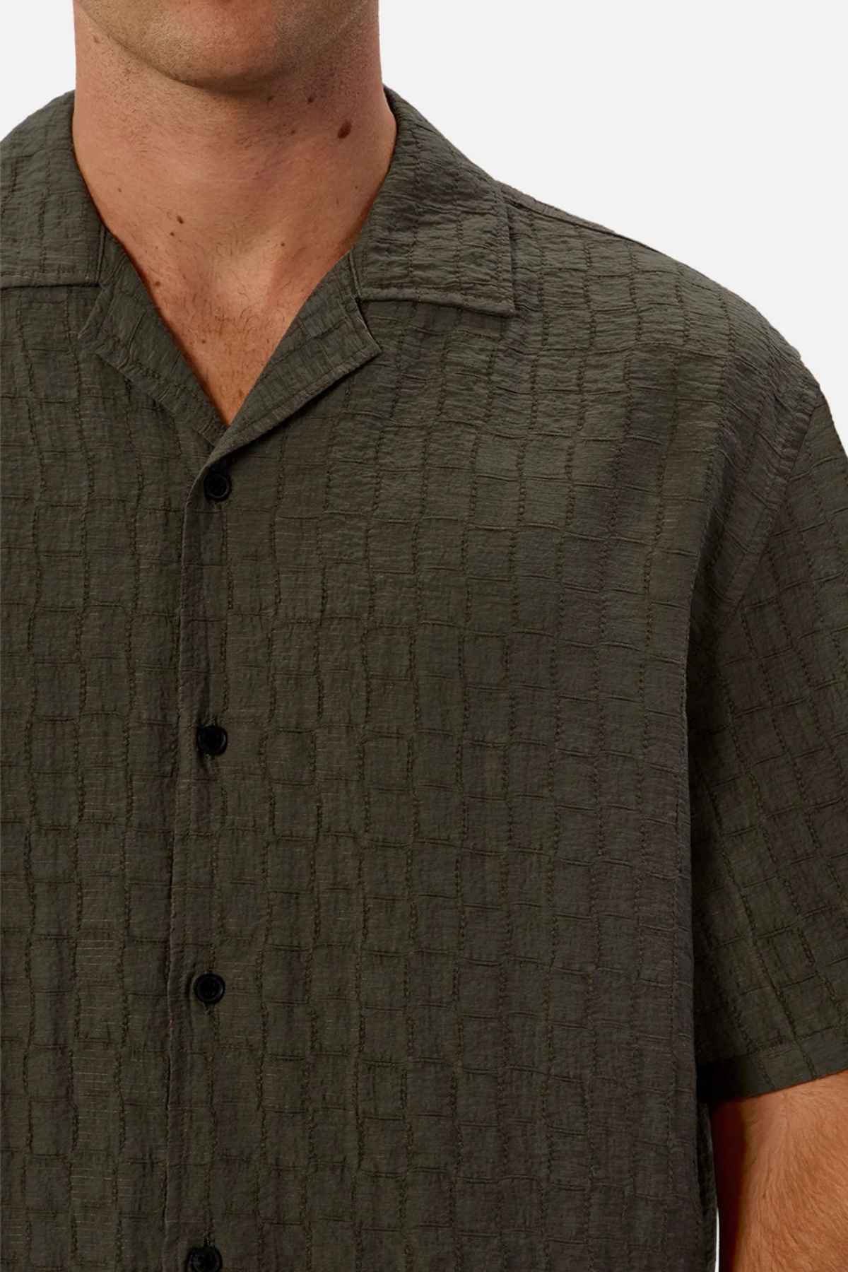 A person wearing a textured, dark green short-sleeve button-up shirt with a camp collar stands against a plain white background. The shirt, an Industrie The New Ortigia Short Sleeve Shirt, features a subtle checkered jacquard pattern with dark buttons. Only the upper torso is visible.