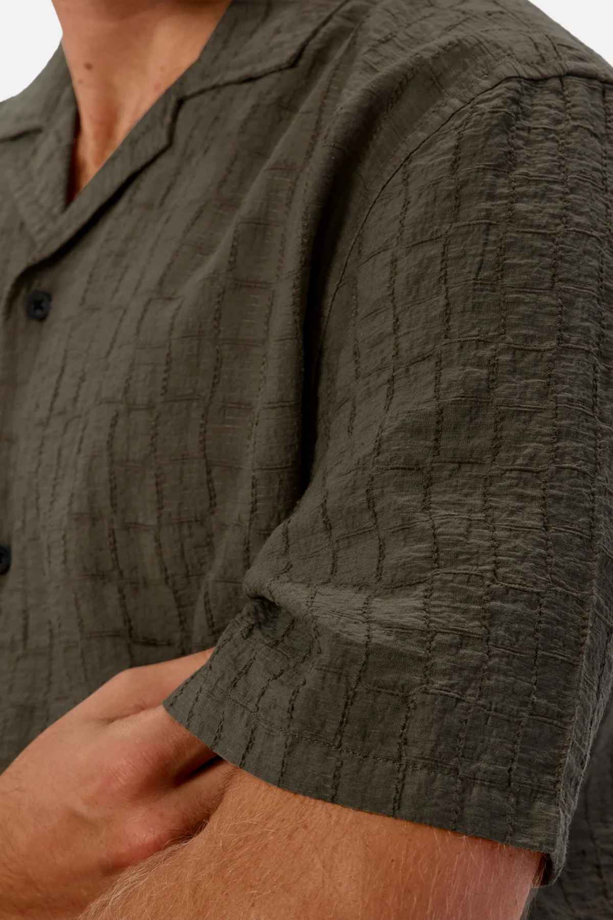 Close-up of a person wearing the Industrie The New Ortigia Short Sleeve Shirt, a textured olive green short-sleeve shirt featuring a checkered jacquard pattern. The person's arms are crossed, and the shirt showcases black buttons and a camp collar. The fabric appears lightweight and casual.