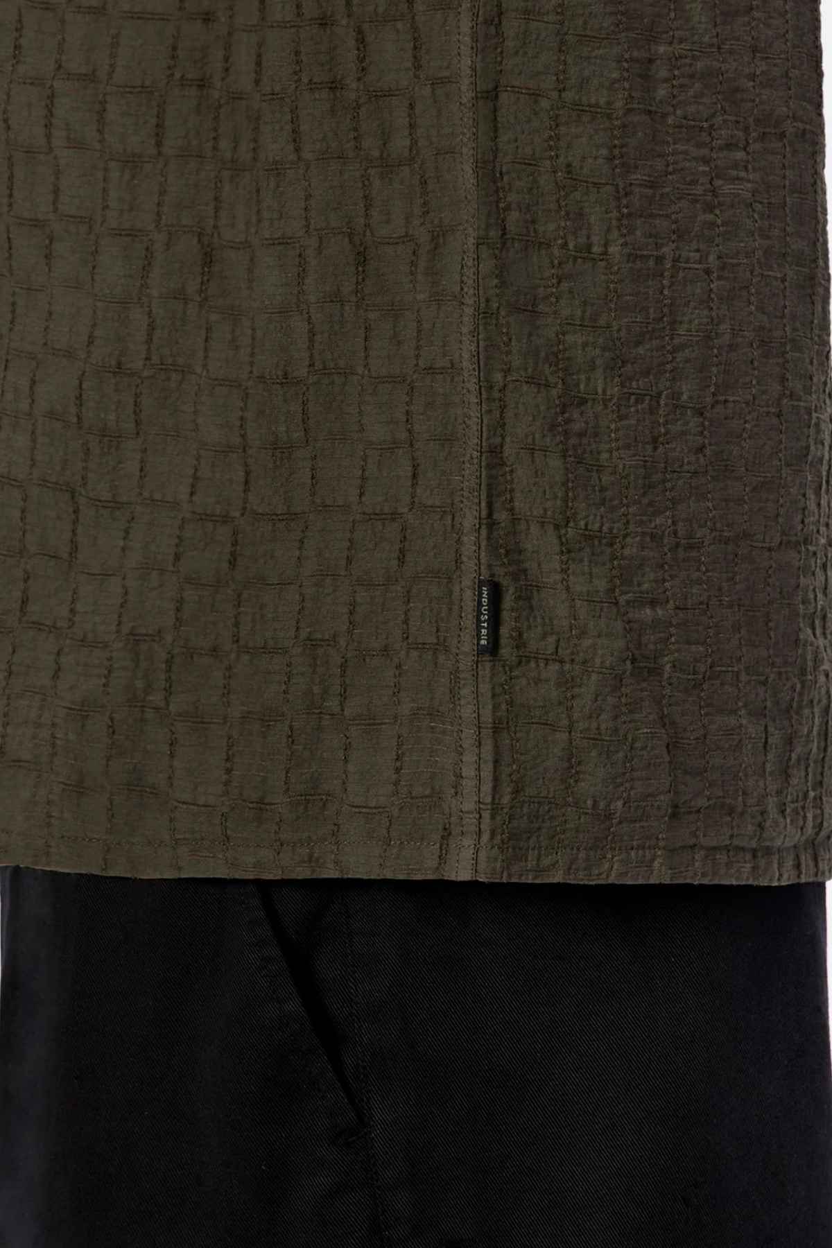 Close-up of a textured, dark green garment with a jacquard pattern, reminiscent of the Industrie The New Ortigia Short Sleeve Shirt. The camp collar design layers over black fabric, and a small tag is visible on the seam.