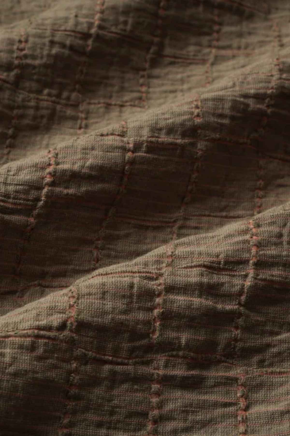 A close-up of the textured fabric with a jacquard pattern showcases a grid-like design, highlighting brown stitching on a deeper brown background. The fabric looks soft and slightly crinkled, similar to the luxurious texture found in an Industrie The New Ortigia Short Sleeve Shirt.