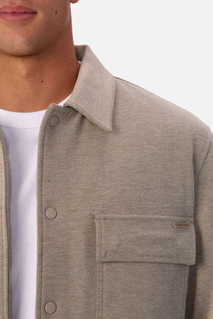 A close-up of someone wearing the Industrie The New Westport Jacket—a light beige textured casual jacket with snap buttons and a front flap pocket—over a white T-shirt, with their face partially visible.