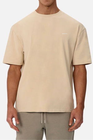 A person stands against a plain background wearing the oversized Industrie The Newport Tee, a loose-fitting 100% cotton shirt with short sleeves and small white text on the upper left chest, paired with taupe drawstring pants.