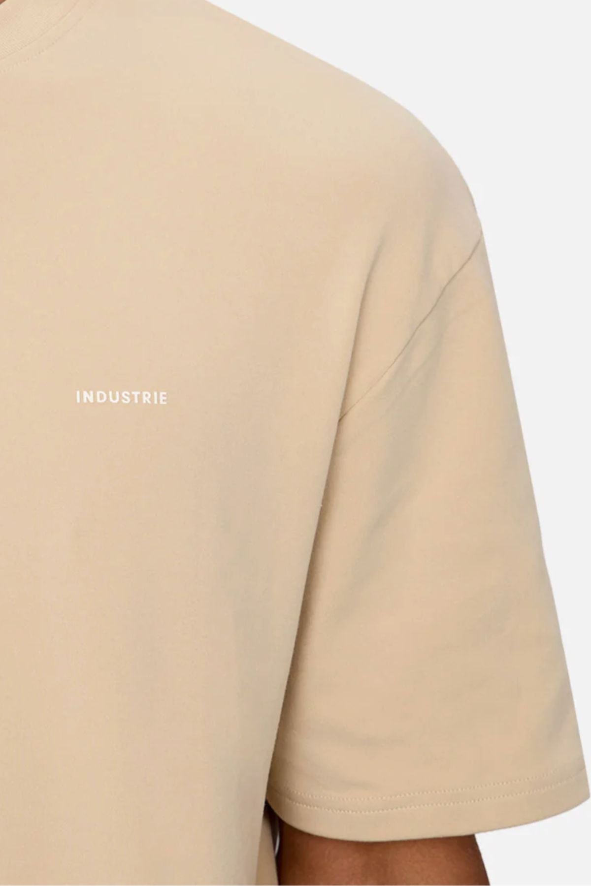 A close-up shows a person wearing the Industrie The Newport Tee in beige, featuring an oversized fit and small white "INDUSTRIE" text on the chest. Crafted from 100% cotton, the T-shirt is set against a plain, light background.