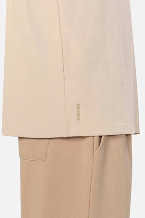 A close-up image of a person wearing a beige, oversized fit shirt and light brown pants. Only the lower part of the Industrie The Newport Long Sleeve Tee and the top of the pants, including the waistband and a pocket, are visible. The fabric of both the shirt and pants appears comfortable and casual.