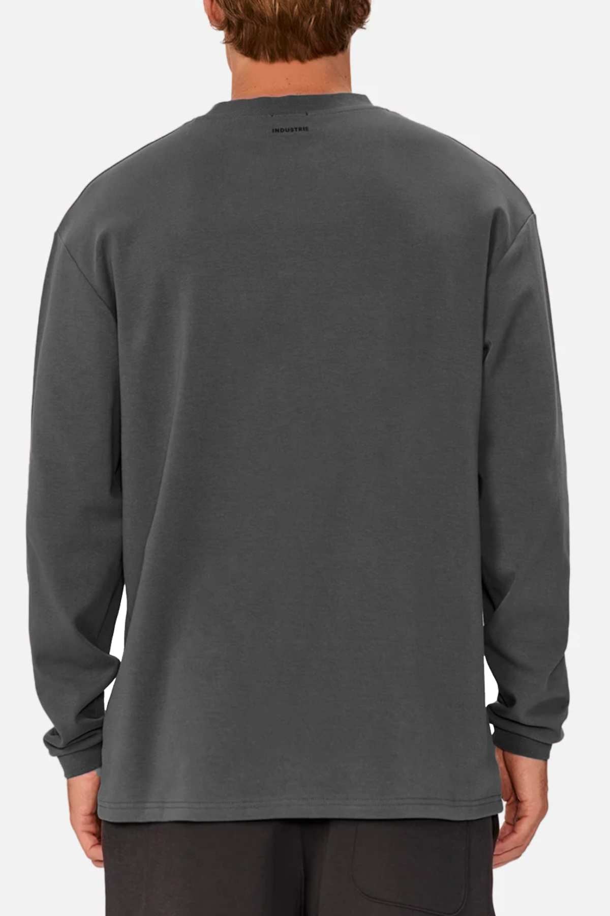 Rear view of a person wearing an oversized Industrie The Newport Long Sleeve Tee in dark gray with a heavyweight finish, paired with dark pants against a light gray background. The shirt has subtle text near the neckline.