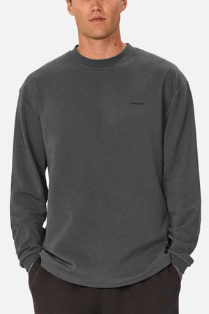A person in a dark gray Industrie The Newport Long Sleeve Tee with subtle chest branding stands against a light gray backdrop, showcasing an effortlessly cool look with its heavy weight finish and oversized fit, as they casually tuck their hands into the pockets of their dark pants.