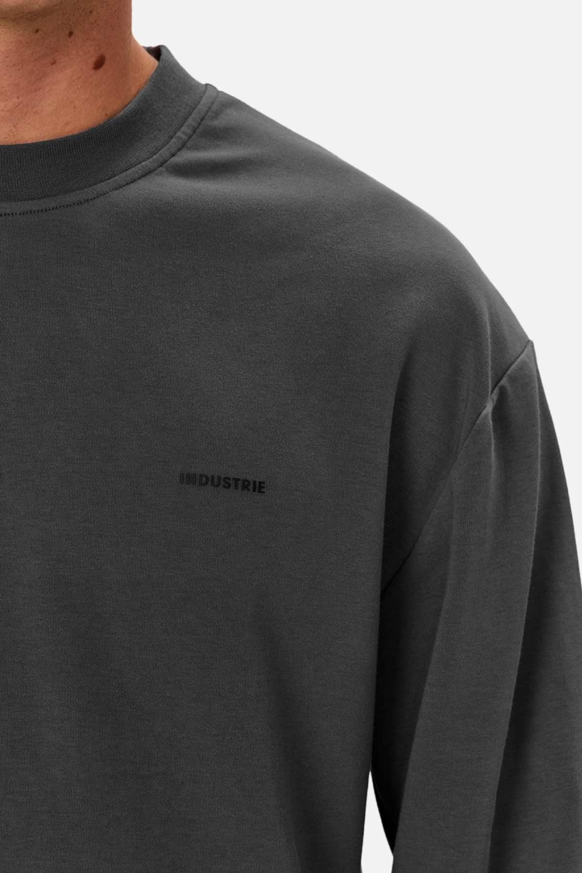 Wearing an oversized, heavy-weight dark gray Industrie The Newport Long Sleeve Tee with "INDUSTRIE" subtly printed on the chest, a person stands against a plain white background, emphasizing the stylish attire while keeping their face unseen.