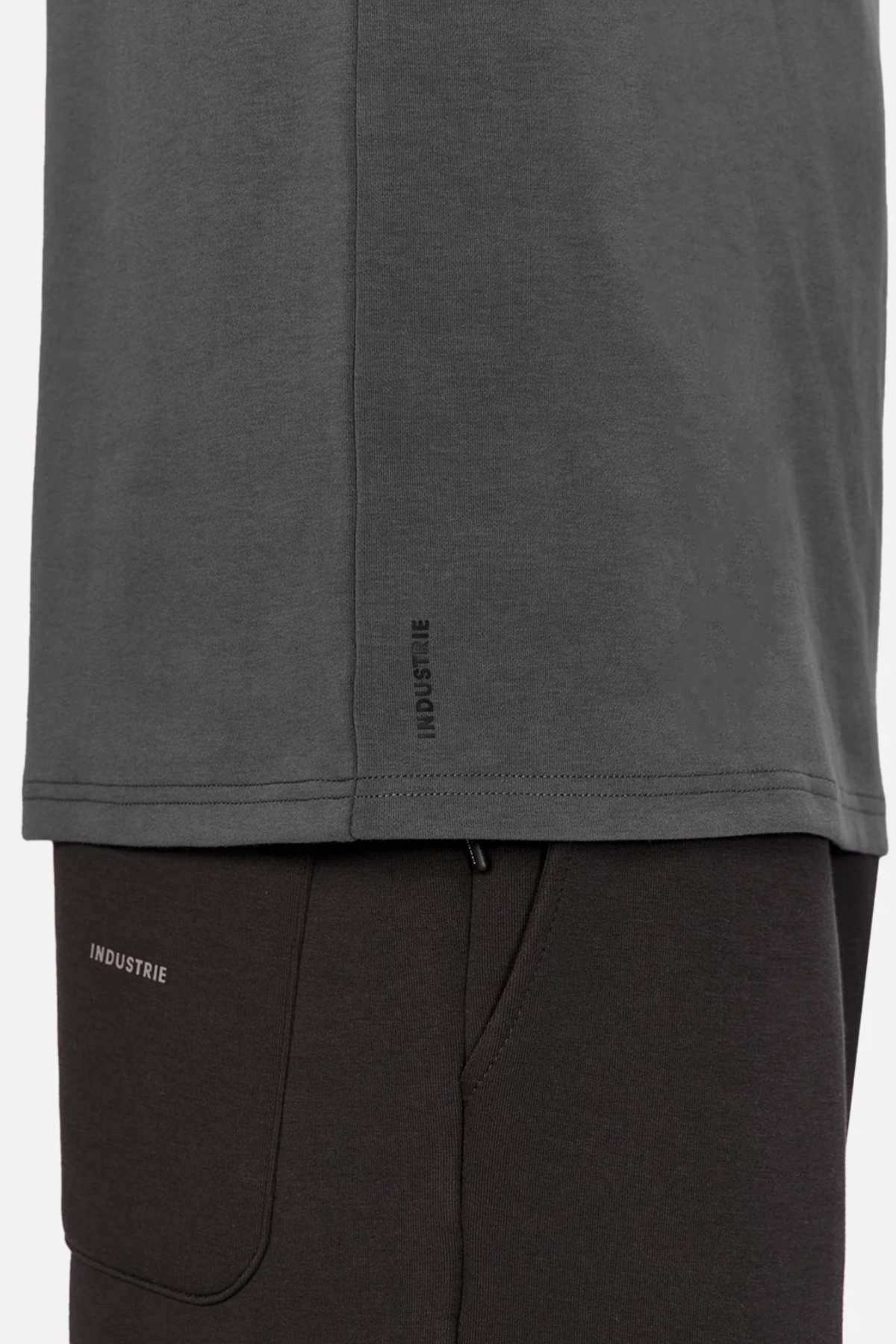 Side view of a person wearing the Industrie The Newport Long Sleeve Tee in dark gray, with "INDUSTRIE" label near the hem, paired with black pants. Focus is on the lower part of the T-shirt and top section of pants.