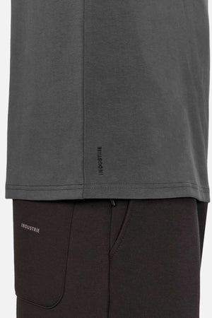 Side view of a person wearing the Industrie The Newport Long Sleeve Tee in dark gray, with "INDUSTRIE" label near the hem, paired with black pants. Focus is on the lower part of the T-shirt and top section of pants.
