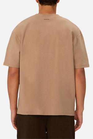 A person is shown from the back wearing an oversized fit beige Industrie The Newport Tee made of 100% cotton, paired with dark pants, against a plain white background.