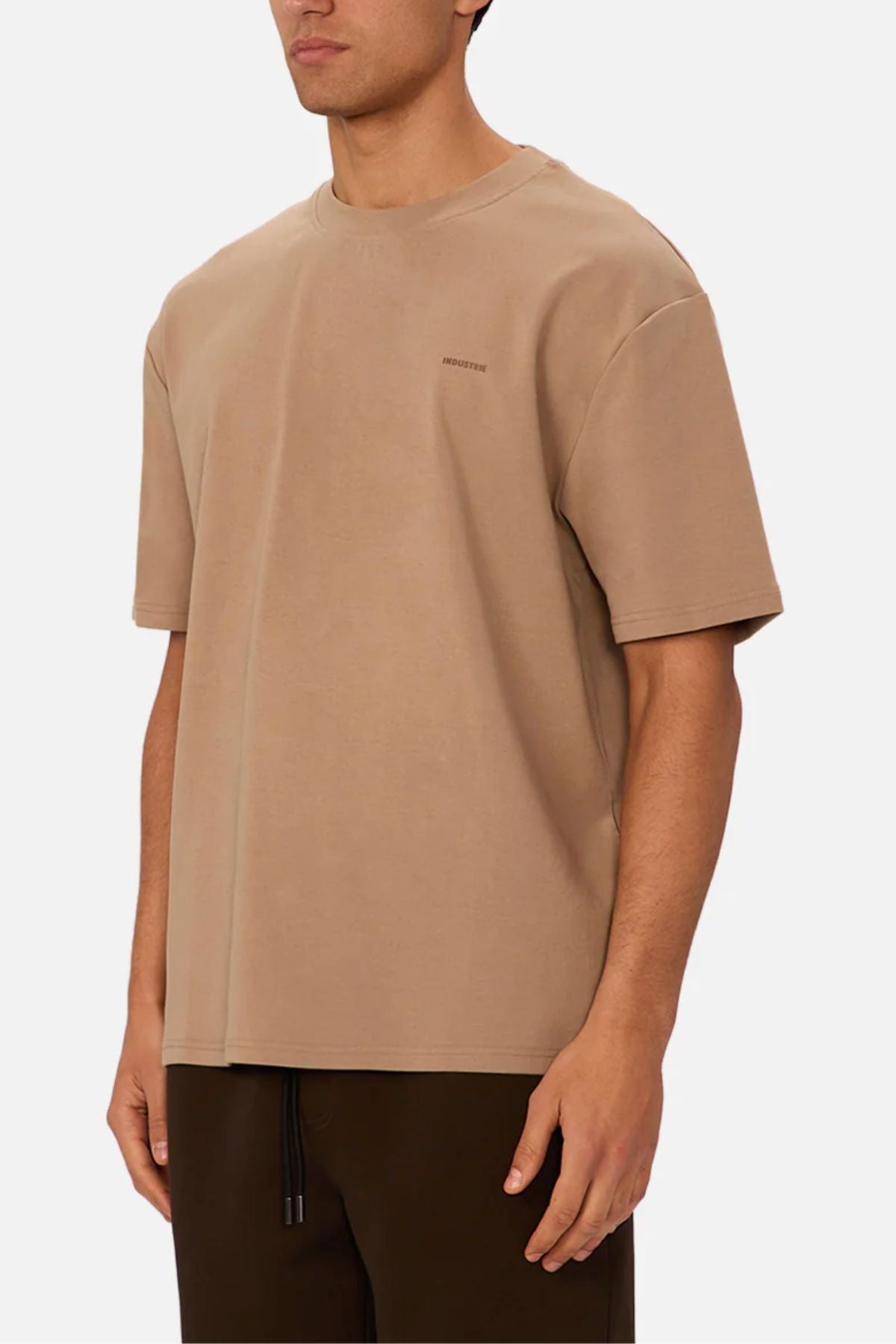 A person wearing the Industrie The Newport Tee in beige, made from 100% cotton, paired with dark brown pants, is standing against a plain white background. Only the upper body and part of their face are visible.
