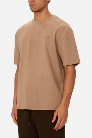 A person wearing the Industrie The Newport Tee in beige, made from 100% cotton, paired with dark brown pants, is standing against a plain white background. Only the upper body and part of their face are visible.