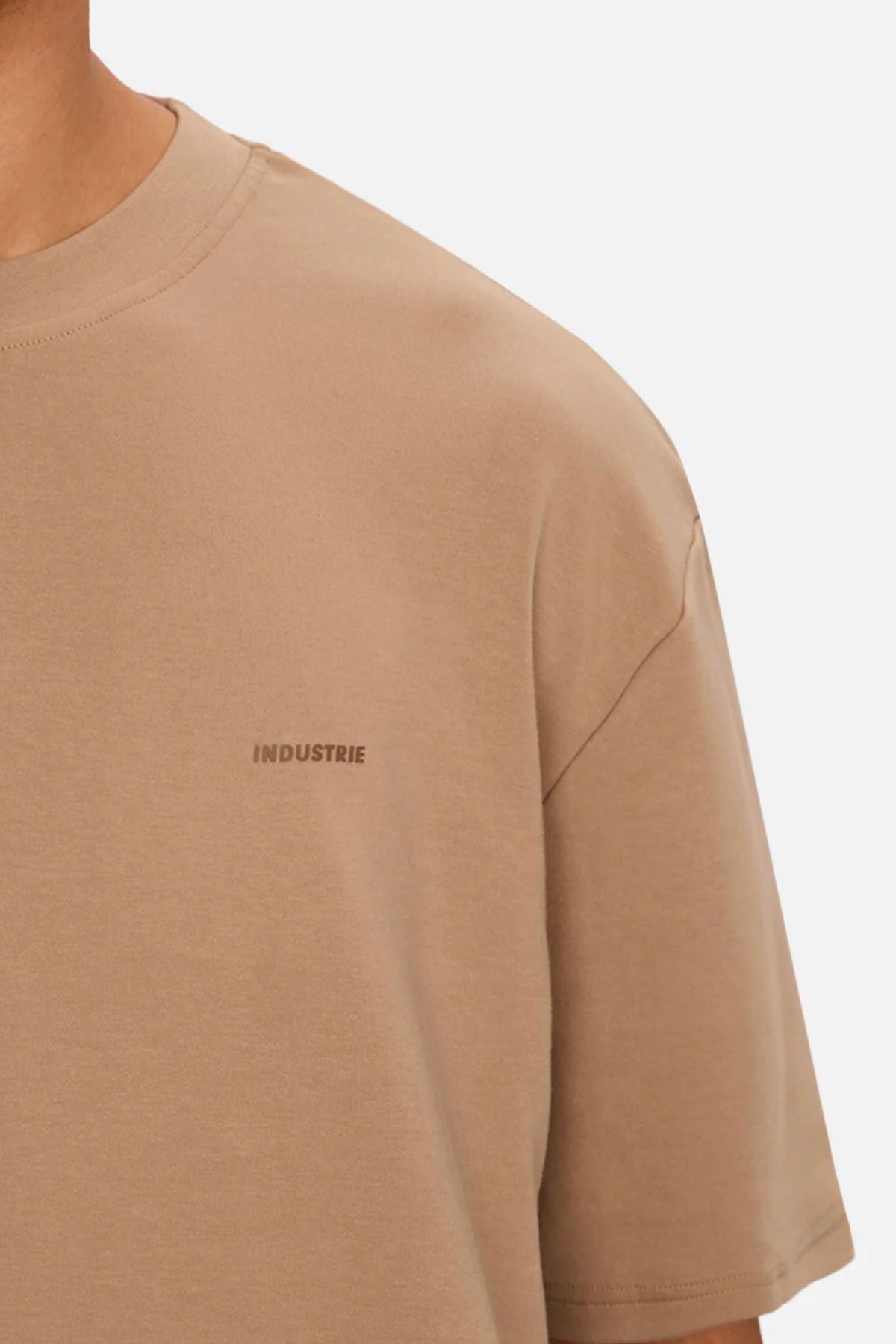 A close-up of someone wearing the Industrie The Newport Tee, an oversized beige 100% cotton t-shirt with a subtle "INDUSTRIE" logo on the chest, set against a plain, light background.