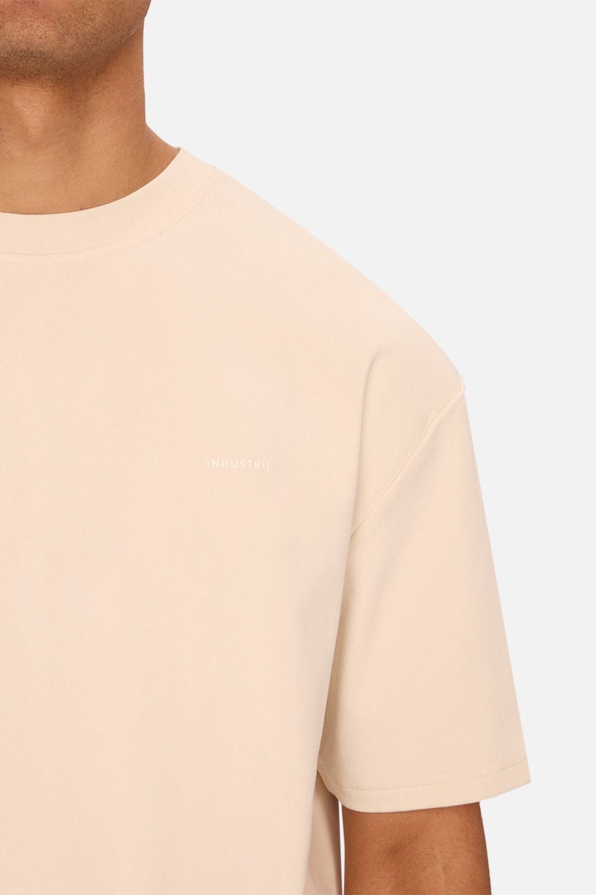 A person wearing the Industrie The Newport Tee, a plain beige oversized t-shirt featuring a small white "INDUSTRIE" print on the left chest, stands against a solid light gray background.