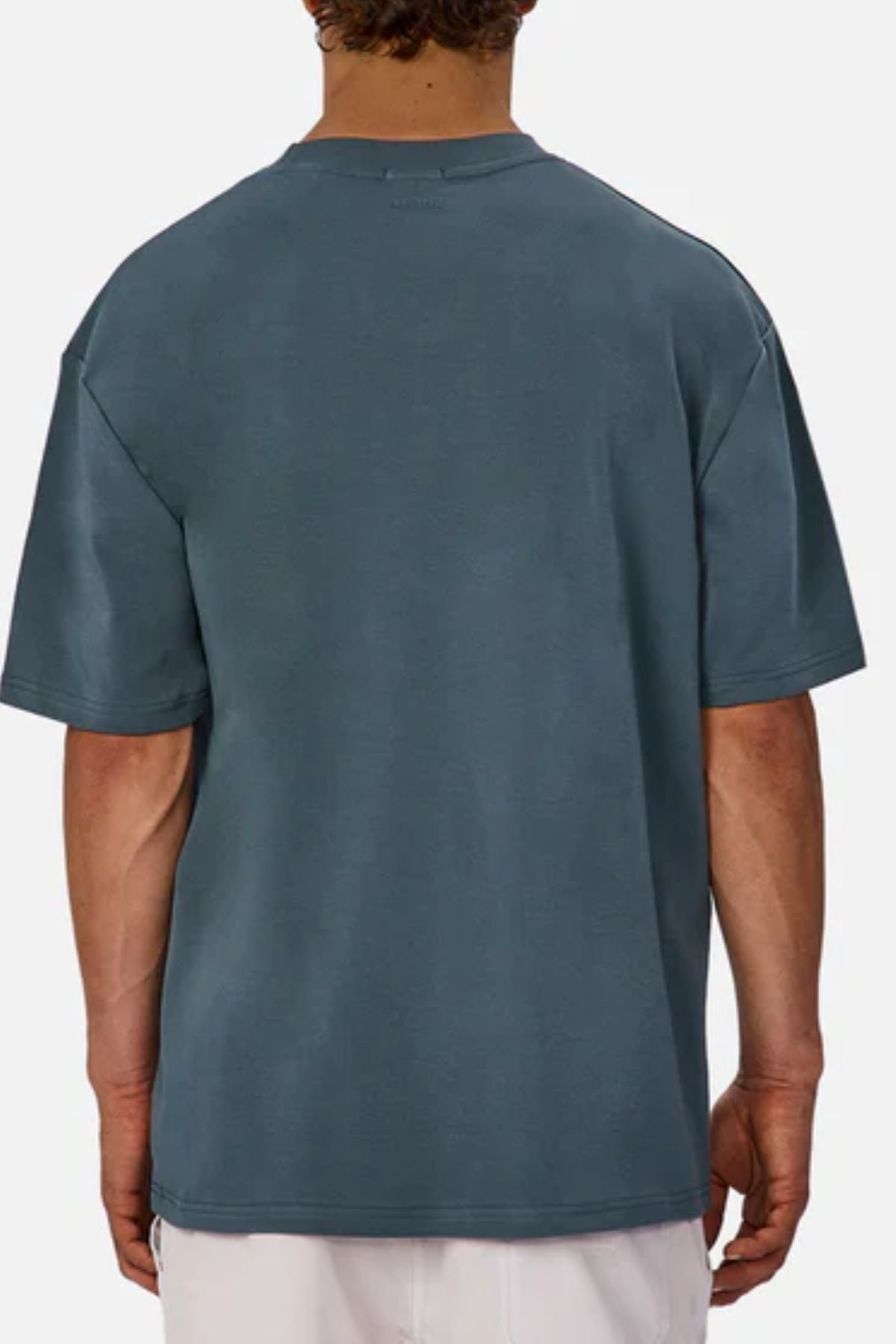A person is seen from behind wearing Industrie The Newport Tee, a relaxed-fit, short-sleeved dark teal heavyweight cotton t-shirt, paired with white pants against a plain light background.