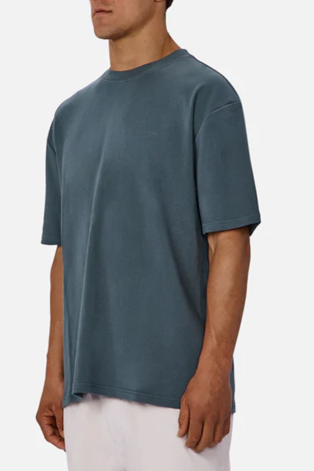 A man stands against a plain, light background wearing the Industrie The Newport Tee in teal with light-colored pants; his face is partially out of frame.
