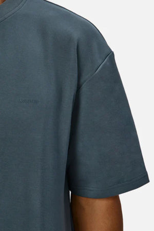 A close-up of someone wearing Industrie The Newport Tee, a dark gray, heavyweight cotton t-shirt with an oversized fit, slightly dropped shoulders, subtle stitching details, and a small logo. Shown against a plain light gray background.