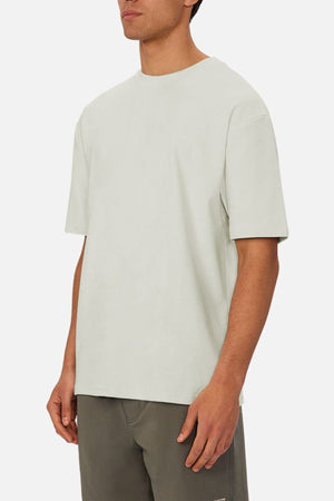A man in an Industrie The Newport Tee, a loose-fitting, heavyweight light gray shirt with an oversized fit, and olive green shorts stands against a plain white background. His face is partially out of view.