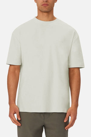 Wearing the Industrie The Newport Tee in plain light gray with an oversized fit and dark pants, a person stands against a white background. The upper body is shown with arms relaxed at their sides.