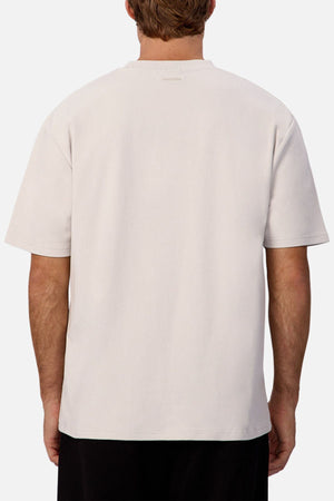 A person is seen from behind wearing an oversized, light-colored Industrie The Newport Tee with dark pants, set against a neutral background.
