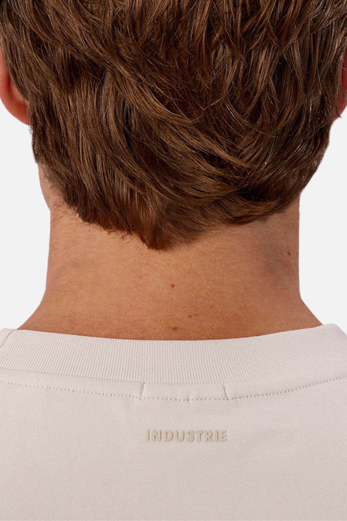 Close-up of the back of a person’s head with short brown hair, wearing an oversized cream-colored Industrie The Newport Tee featuring "INDUSTRIE" printed below the collar.