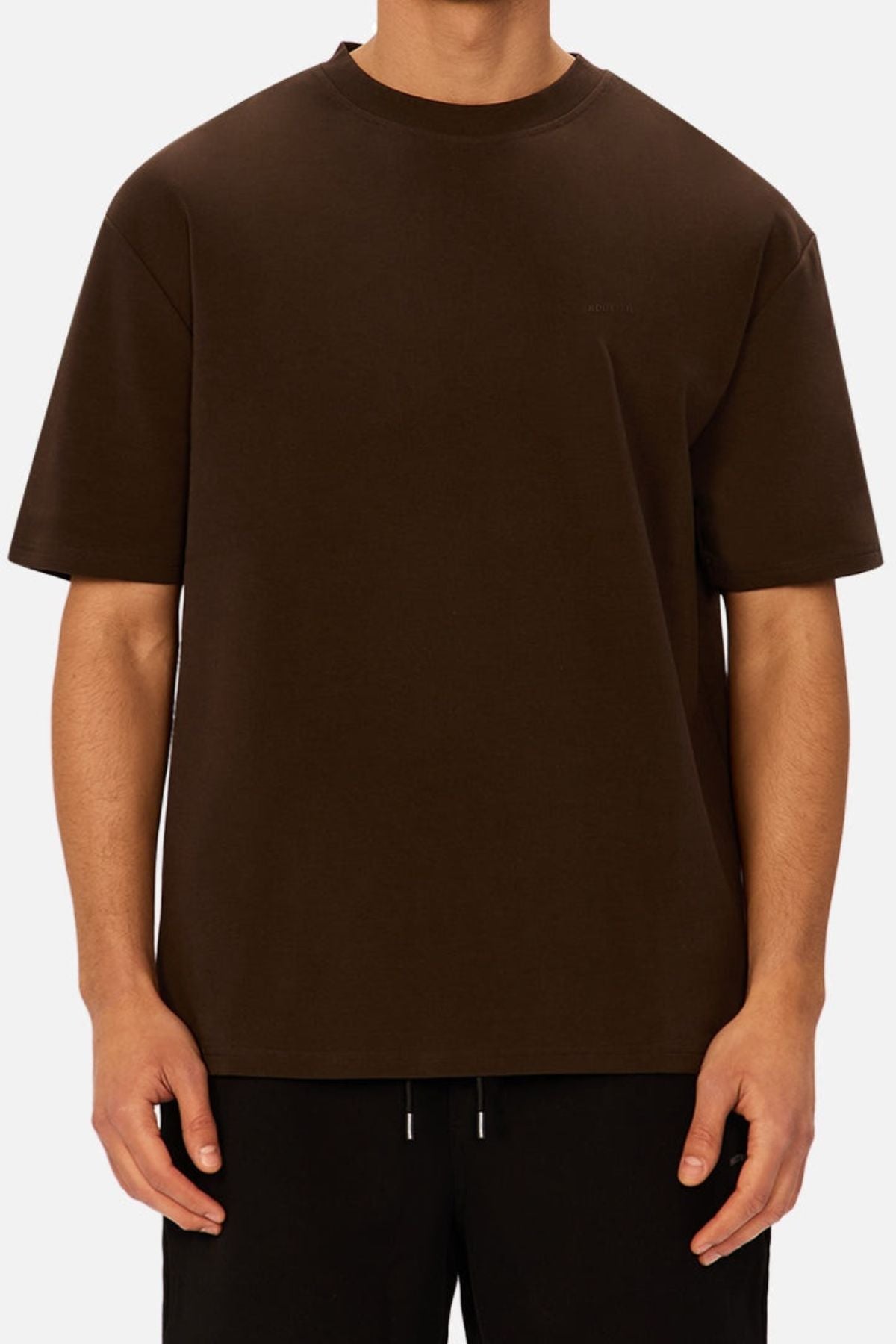 A person wearing an oversized, plain dark brown Industrie The Newport Tee and black pants stands against a light gray background, with their head not visible in the image.
