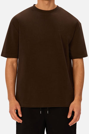 A person wearing an oversized, plain dark brown Industrie The Newport Tee and black pants stands against a light gray background, with their head not visible in the image.