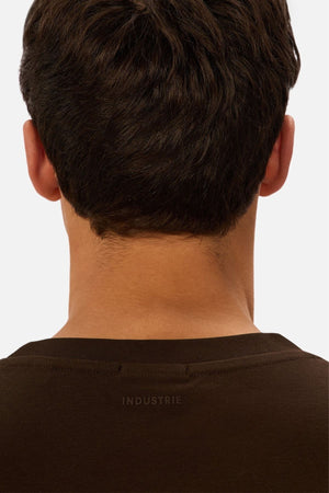 The image displays the back of a man's head and neck with short dark hair, wearing the Industrie The Newport Tee in dark brown, oversized fit, and "INDUSTRIE" printed near the collar.
